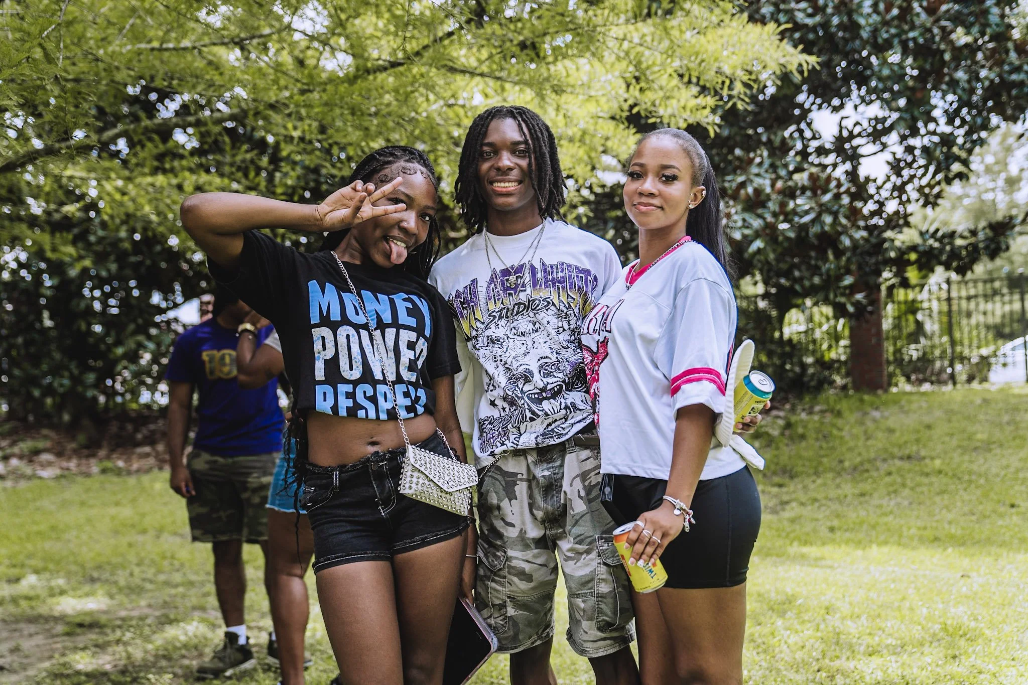 Three young women posing together outdoors on a grassy area with trees in the background, smiling and making playful gestures.