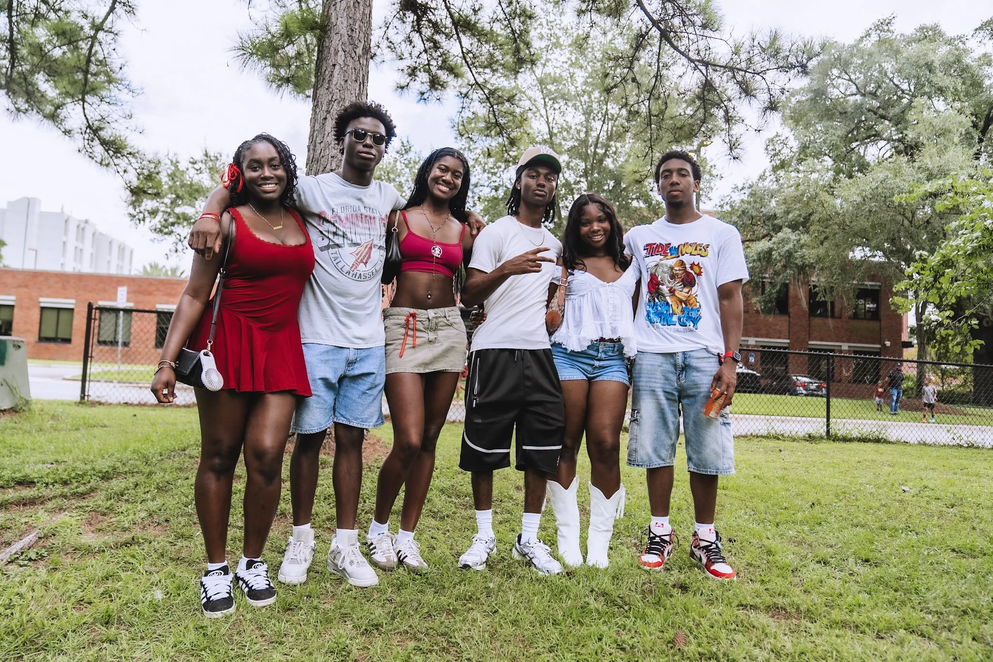Group of six young adults standing outdoors in a park, posing for a photo under a large tree.