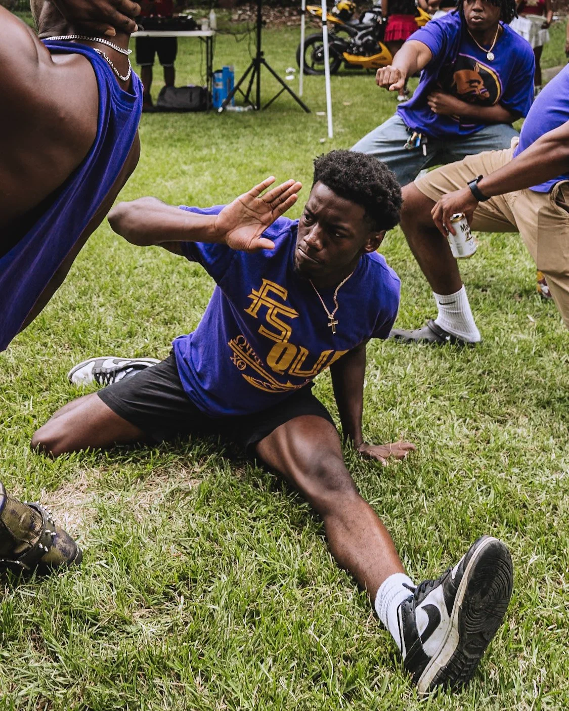 A young man kneels on the grass, leaning back on his left hand, with his right hand raised for a high five with another person outside the frame. He looks serious and is wearing a purple shirt, black shorts, and Nike sneakers.