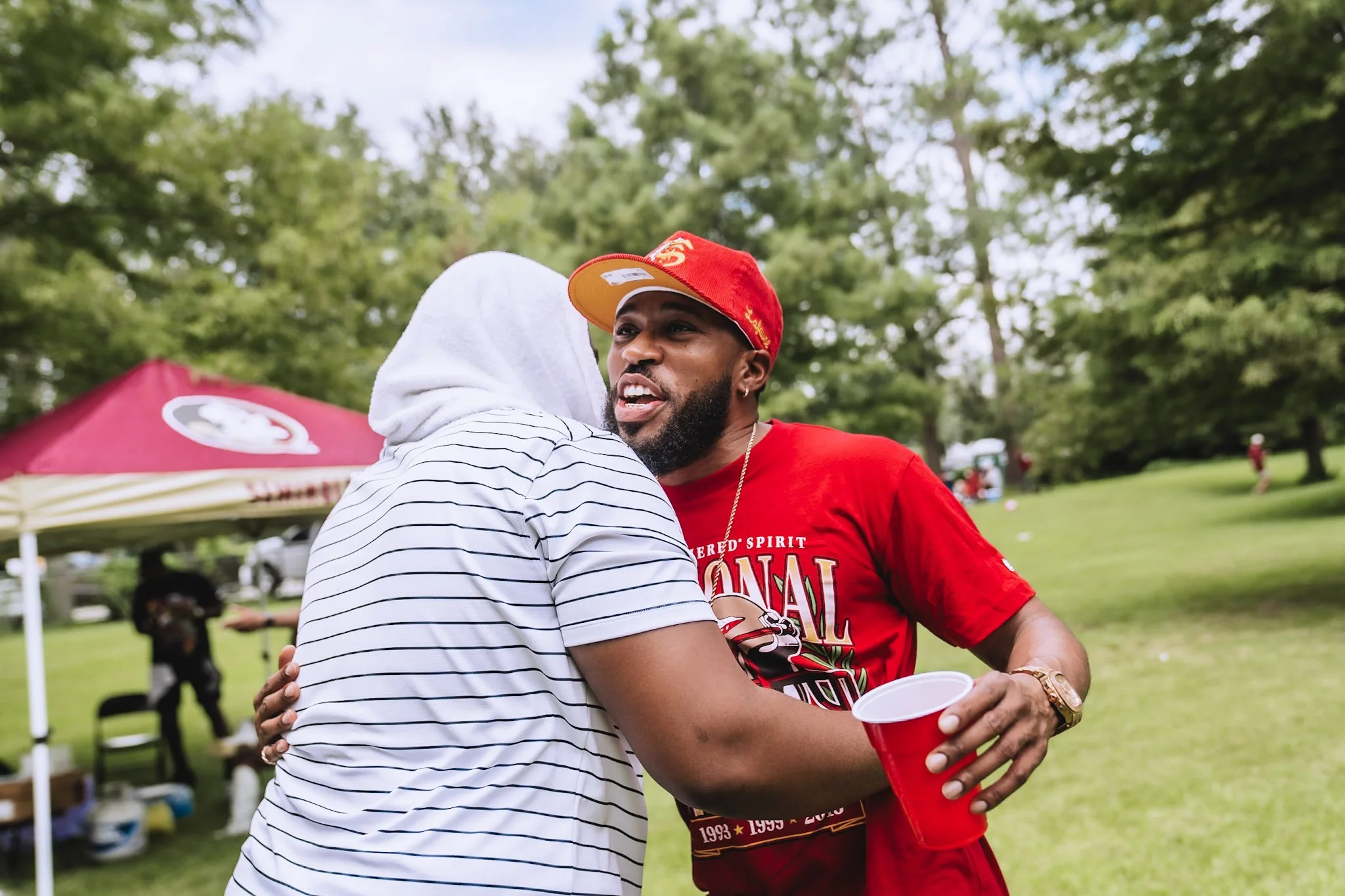 Two men hugging outdoors at a gathering or party, with a red canopy tent in the background, one man wearing a hoodie and the other in a red shirt and cap holding a red cup.