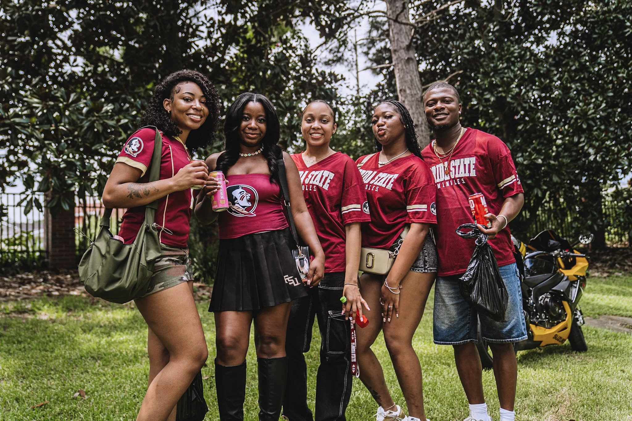 Group of five young adults, four women and one man, wearing Florida State football jerseys and casual clothing, standing outdoors in front of trees, smiling for a photo. They are holding drinks and shopping bags, with a yellow motorcycle in the backg