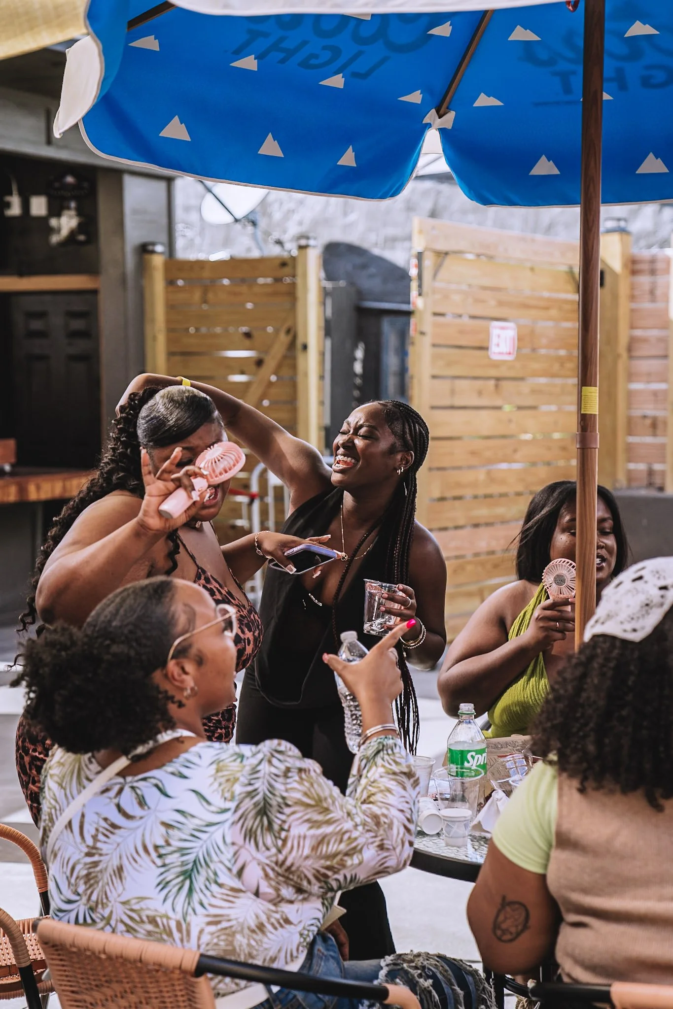 Group of women enjoying a casual outdoor gathering under a blue umbrella, sitting around a table with drinks.