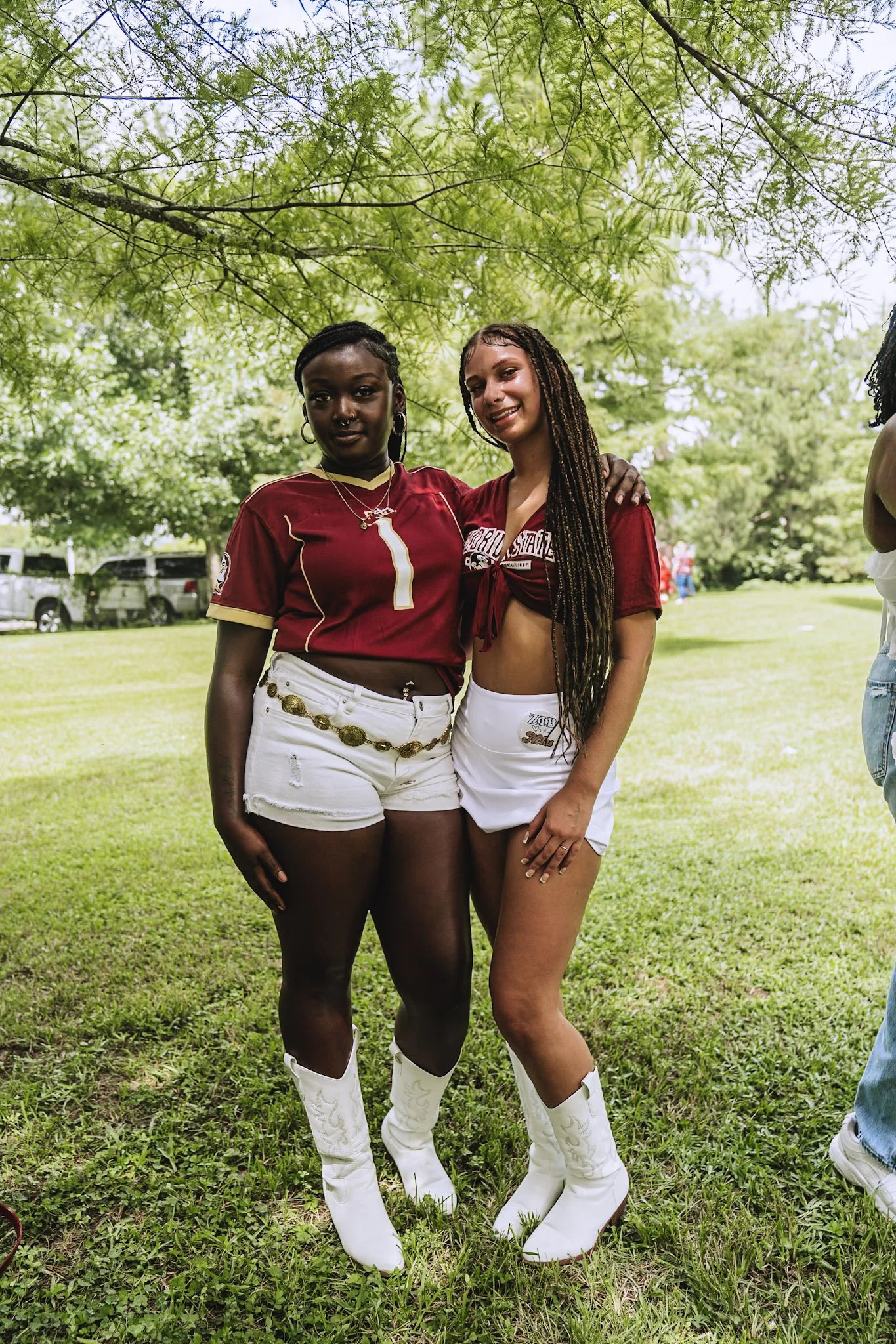Two young women standing close together outdoors under green tree branches, smiling, with parked cars in the background. They are wearing university-themed clothing, white shorts, and white cowboy boots.