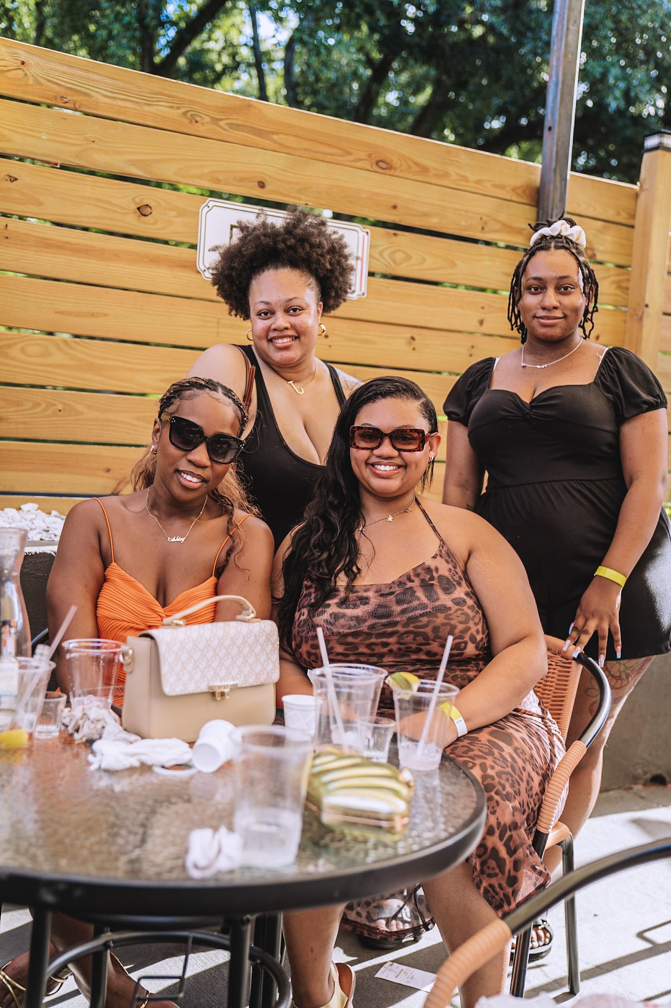 Five women, some wearing sunglasses, sitting and standing around outdoor table with drinks and food, smiling at the camera, with a wooden fence and trees in the background.