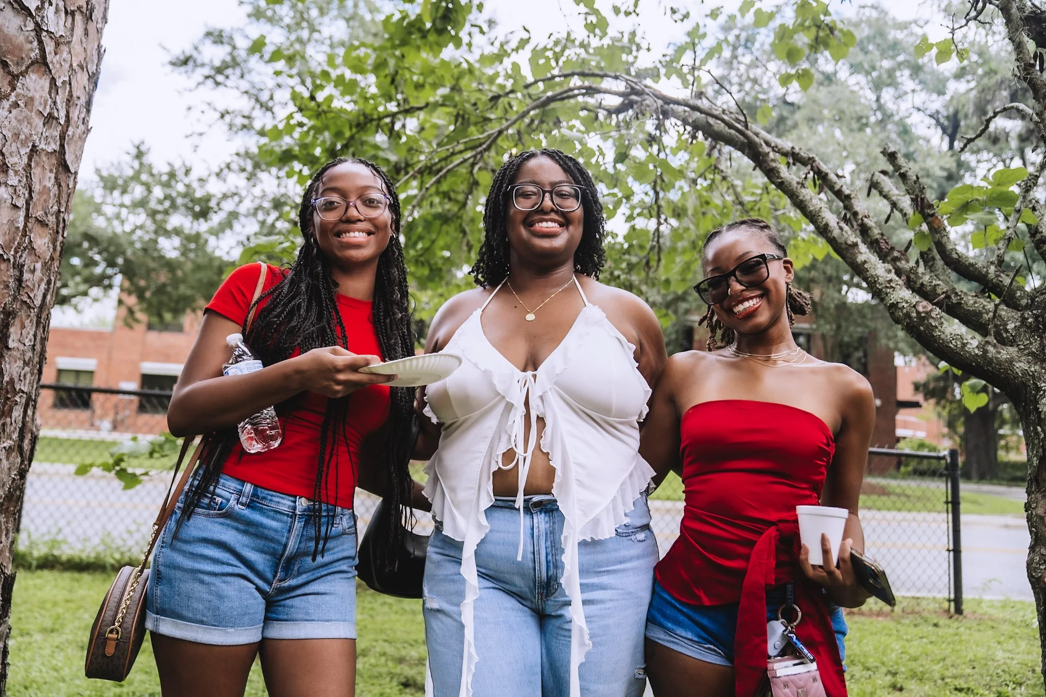 Three young women standing outdoors under a tree, smiling at the camera. They are dressed casually in summer clothing, and appear to be enjoying a social gathering or picnic.