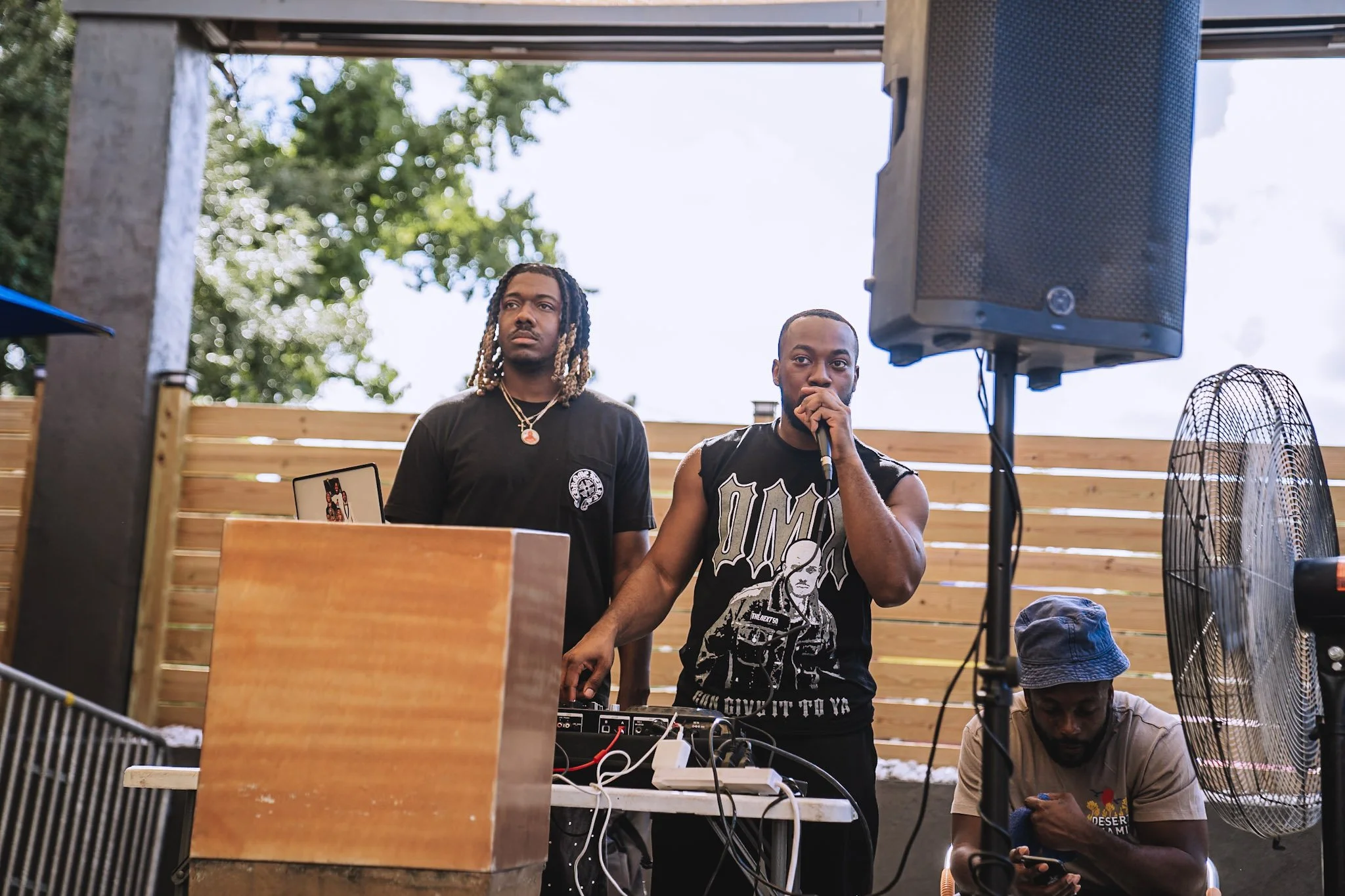 Two men are behind DJ equipment, one is speaking into a microphone, and a third person is sitting on the ground looking at their phone. They are outdoors with a wooden fence in the background.
