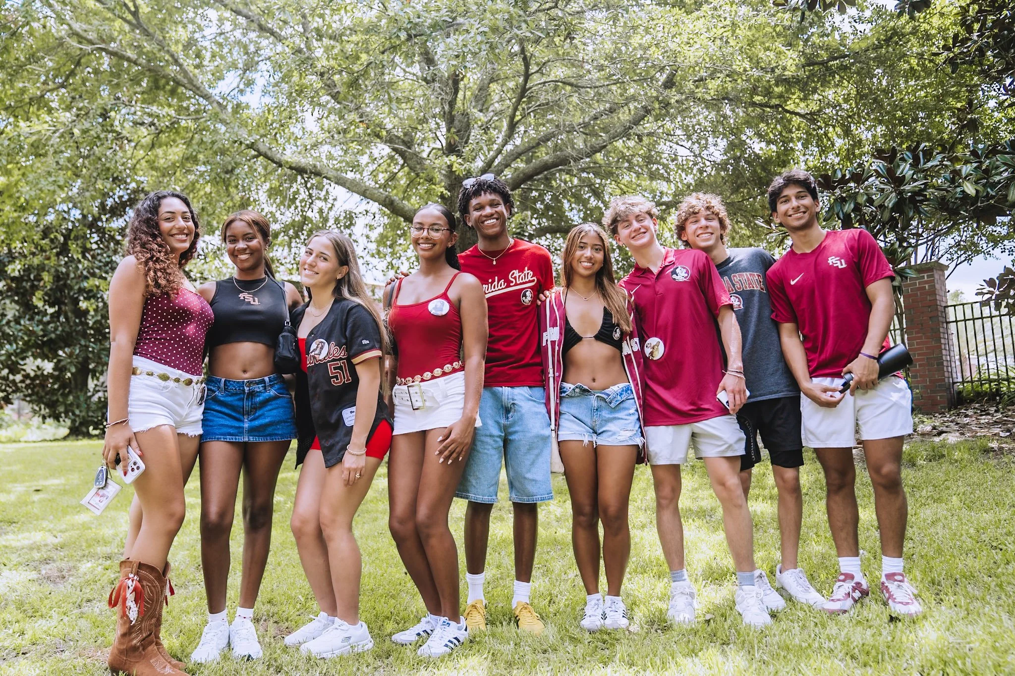 Group of nine diverse young adults standing outdoors on grass, smiling, with trees and a brick fence in the background.