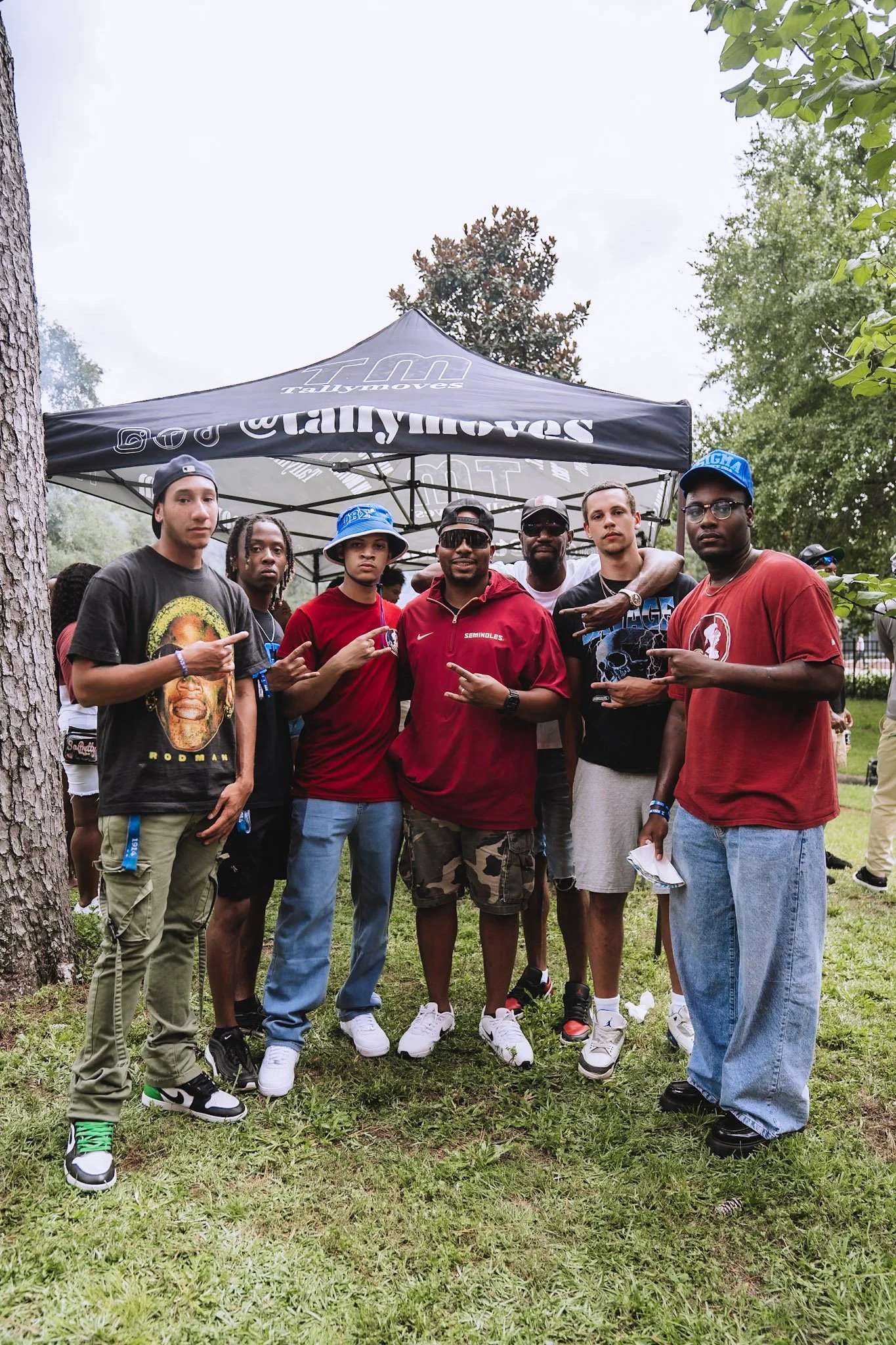 Group of young men posing outdoors under a canopy, some making peace signs, in a grassy park-like setting with trees.