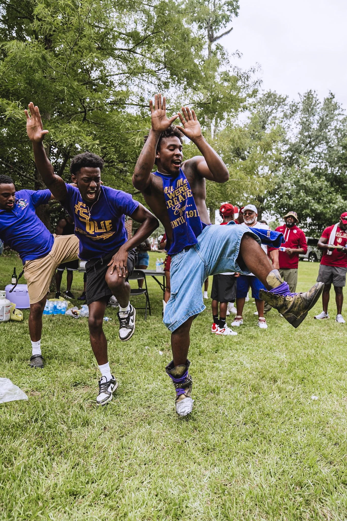 Group of men wearing fraternity shirts participating in a lively outdoor celebration or dance event in a park, with others watching in the background.
