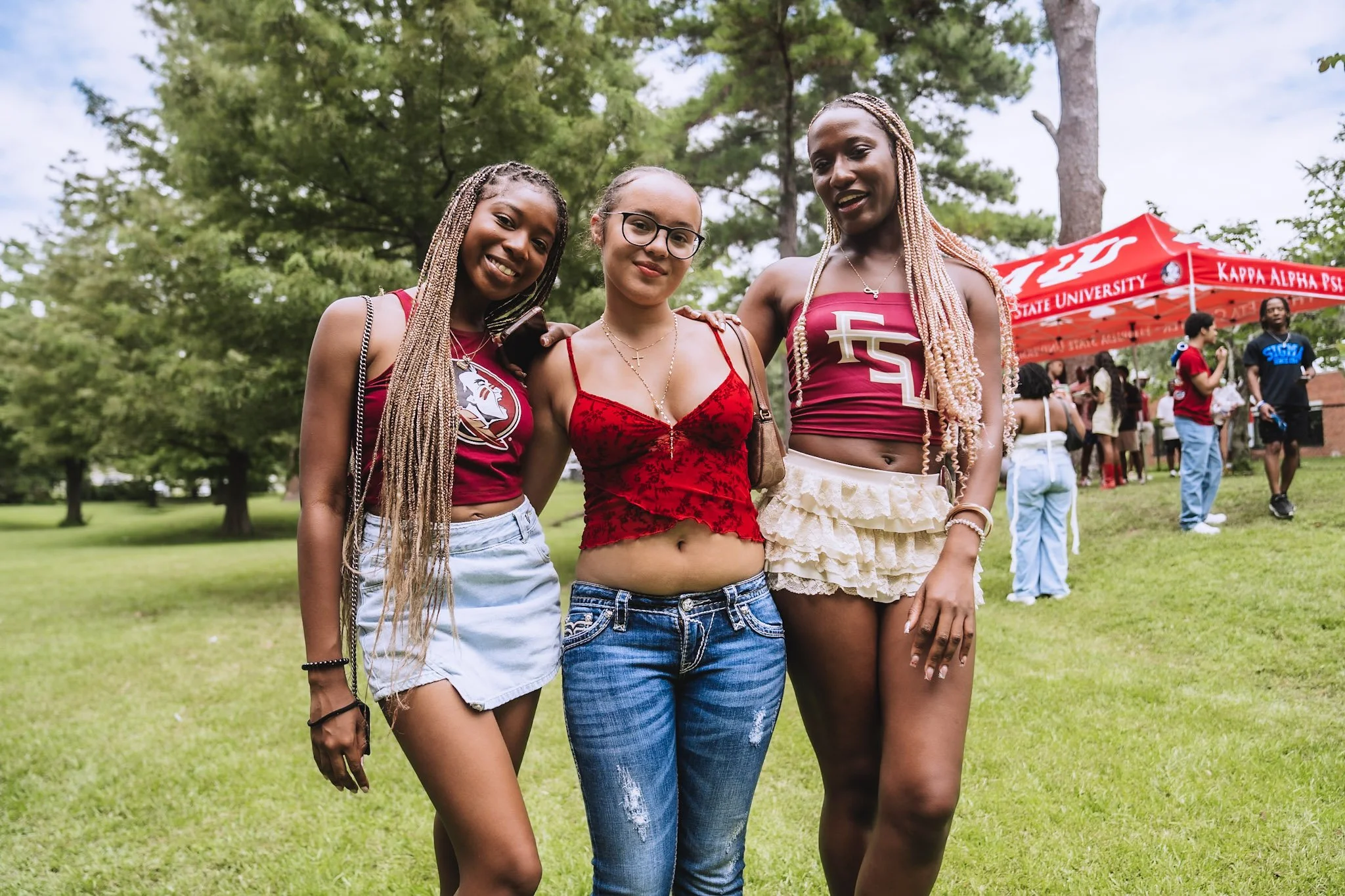 Three young women standing outdoors on a grassy field, smiling, with trees and a red university tent in the background.