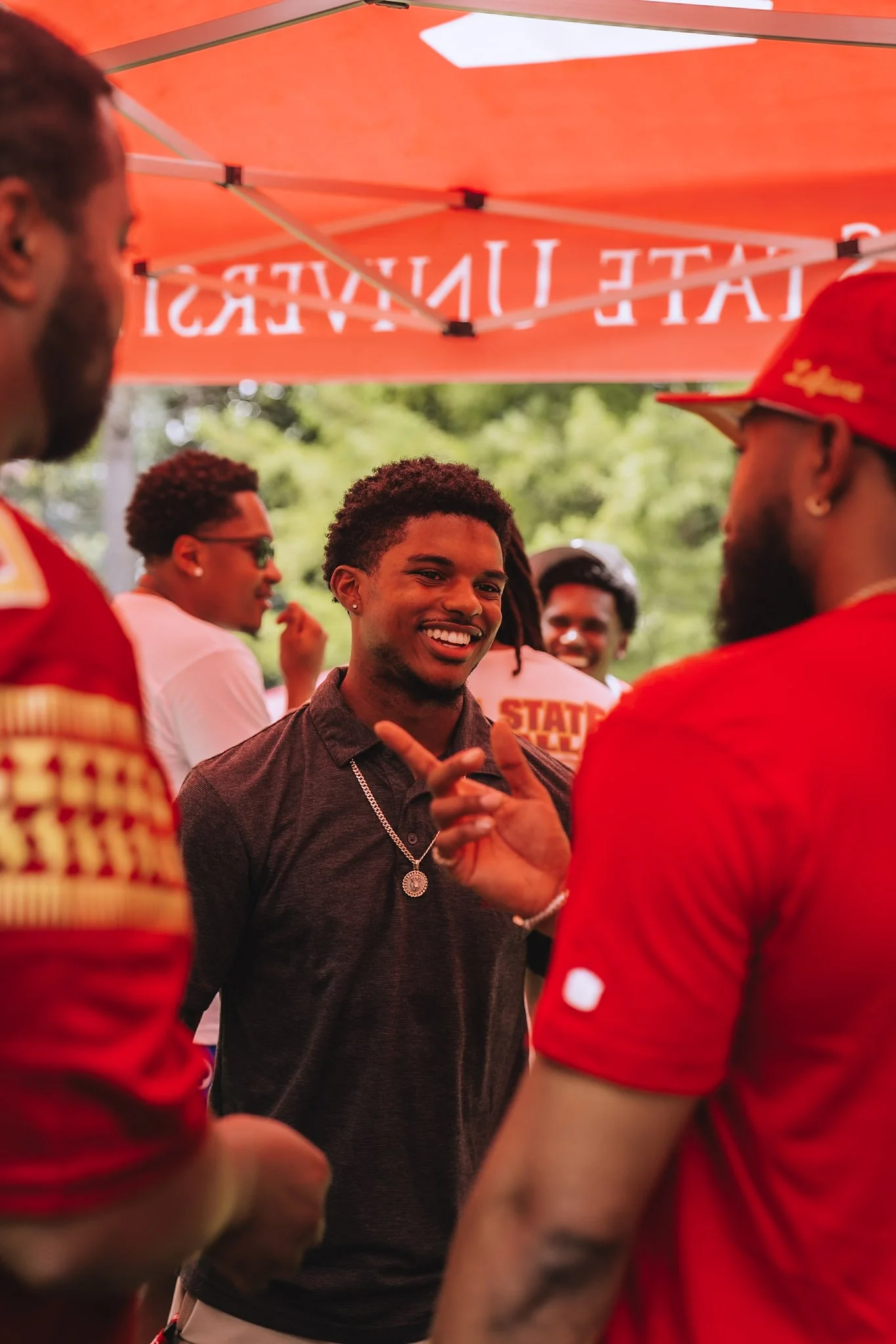 A group of young men, some wearing red shirts, are gathered under a red canopy. One young man in a dark shirt and chain is smiling and talking to another man in a red cap.