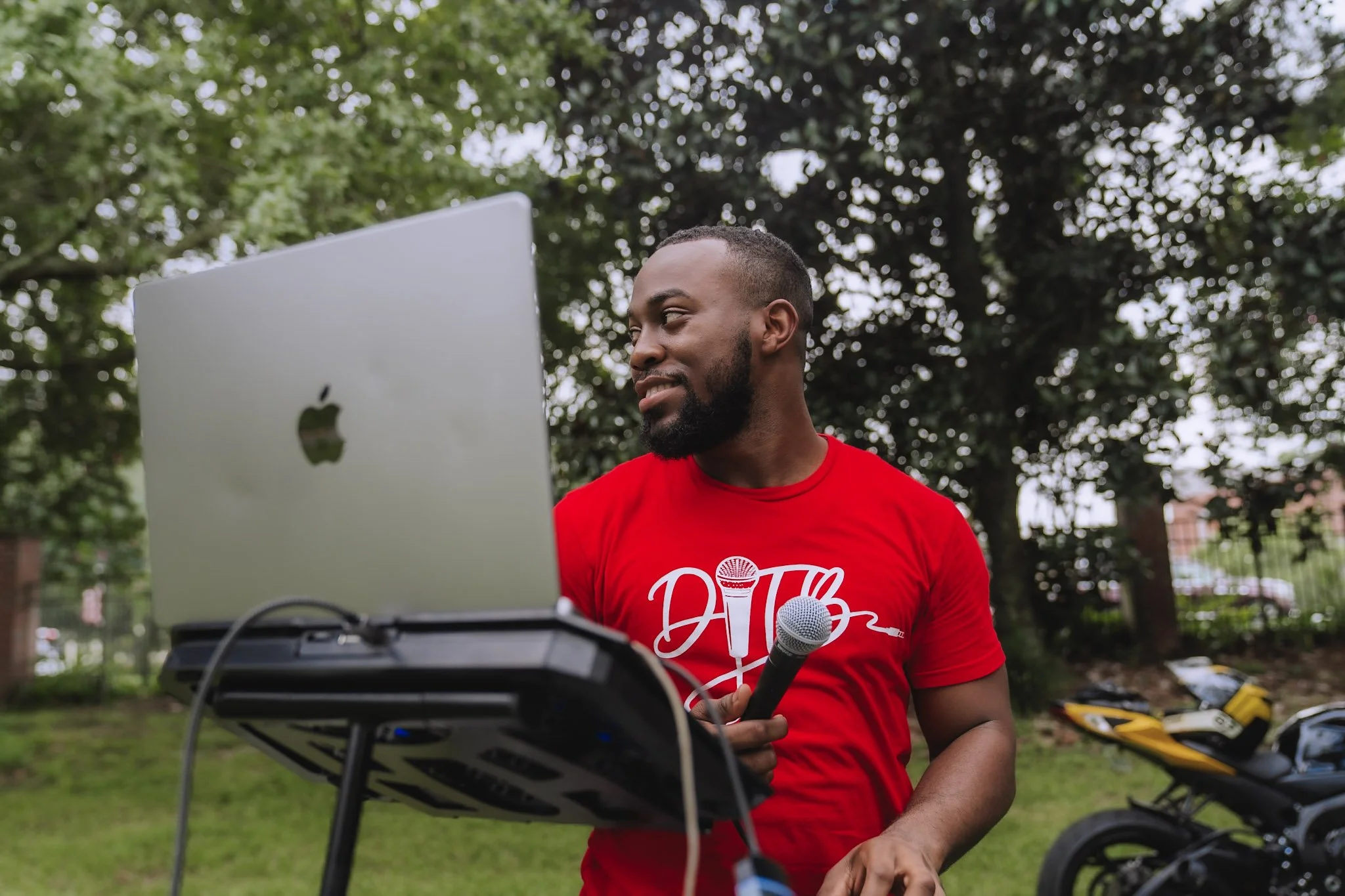 A man in a red shirt holding a microphone, standing outdoors in front of a laptop on a stand, with trees and motorcycles in the background.