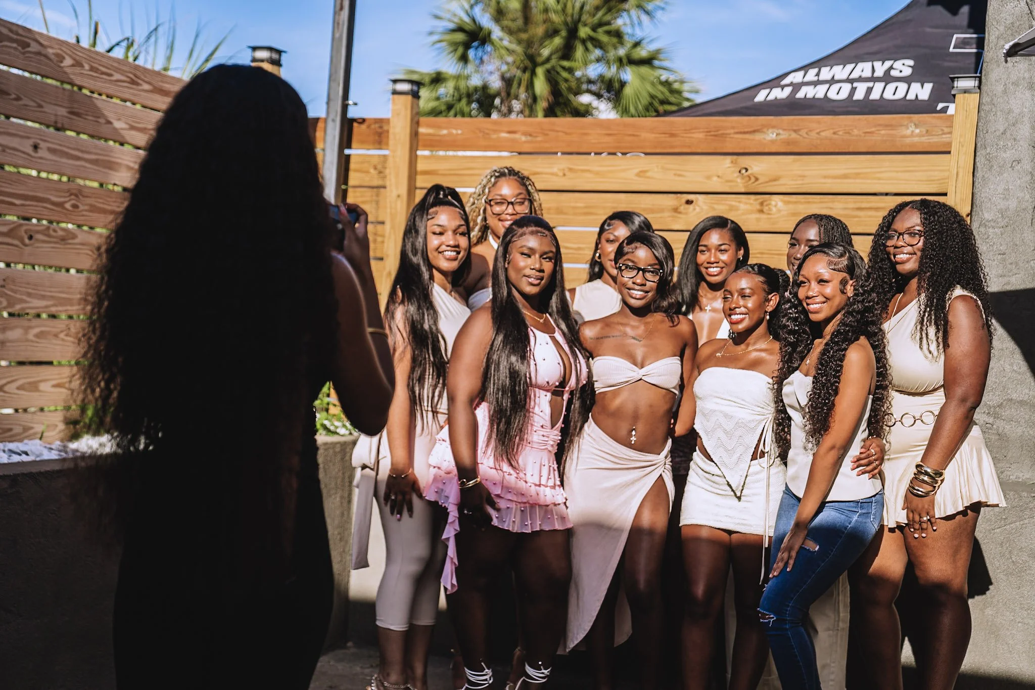 A woman takes a photo of a group of women posing together outdoors, with a wooden fence and a tent with words "Always in Motion" in the background.