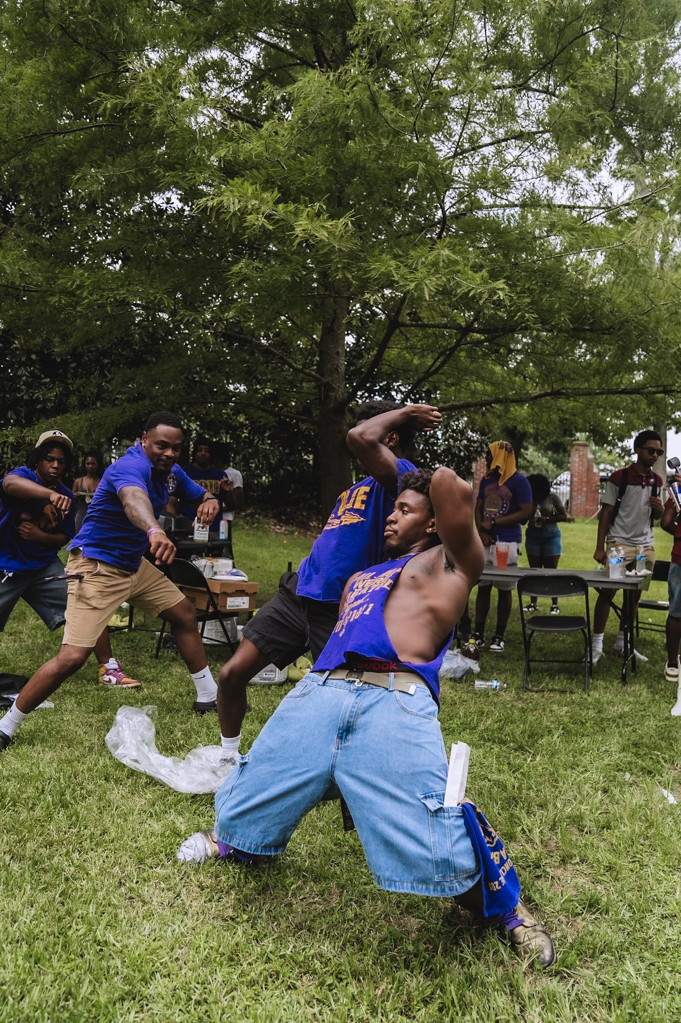 A group of young men strolling outdoors on a grassy area, with some standing and others kneeling or sitting near a table with drinks and supplies.