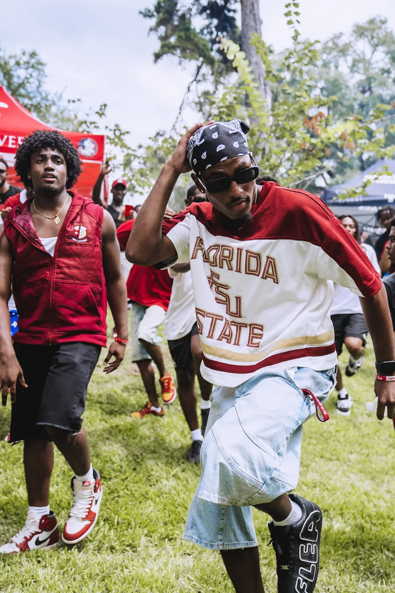 A group of young people dancing and enjoying an outdoor event on a grassy field, with trees and marquees in the background.