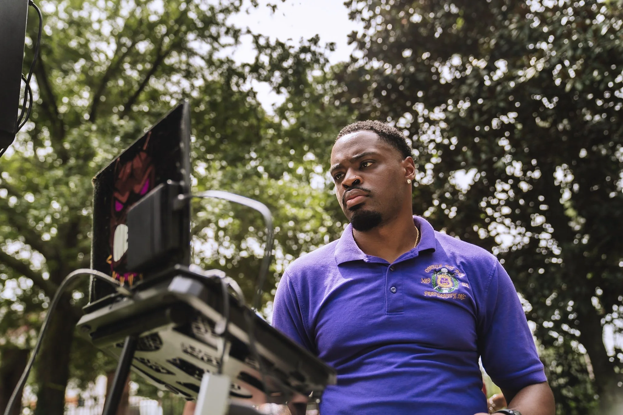 A man in a purple polo shirt with a logo on it stands outdoors in front of trees, looking at a computer setup with wires and screens.