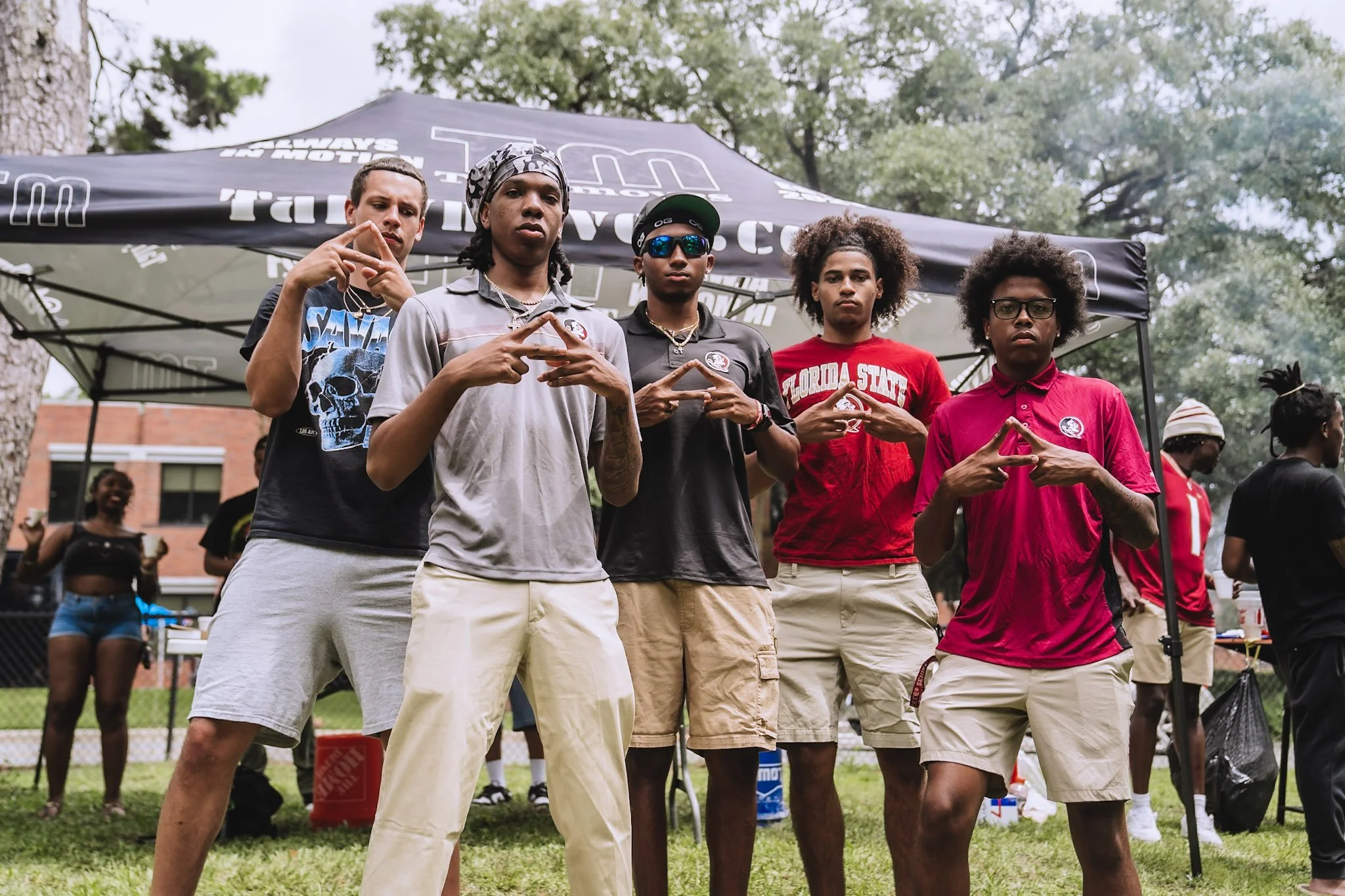 Five young men standing outdoors in front of a black canopy tent with white text, making triangular hand gestures, with trees and other people in the background.