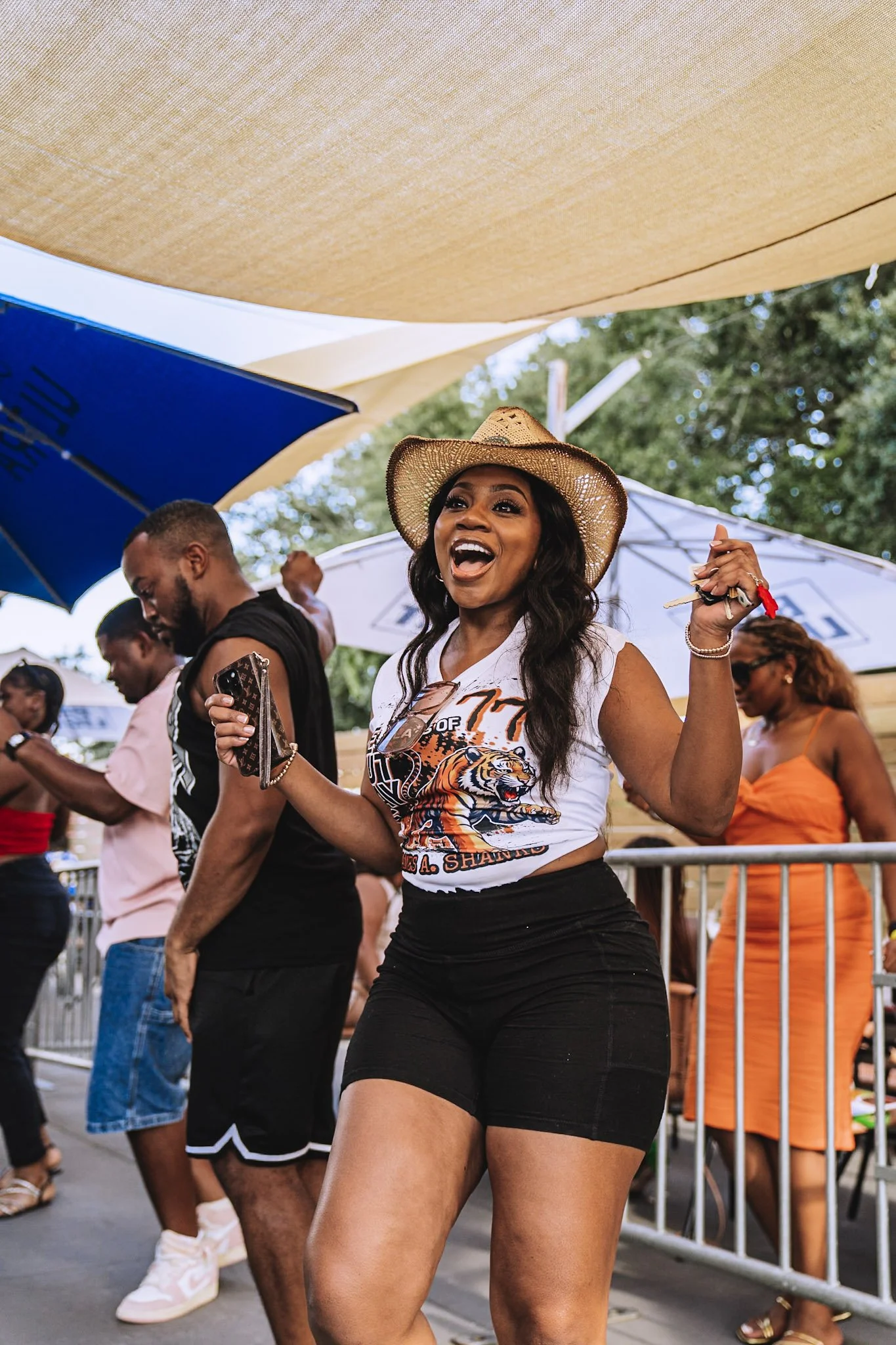 A woman wearing a wide-brimmed hat, a white graphic T-shirt, and black shorts, smiling and holding a drink at an outdoor event under a tan canopy.