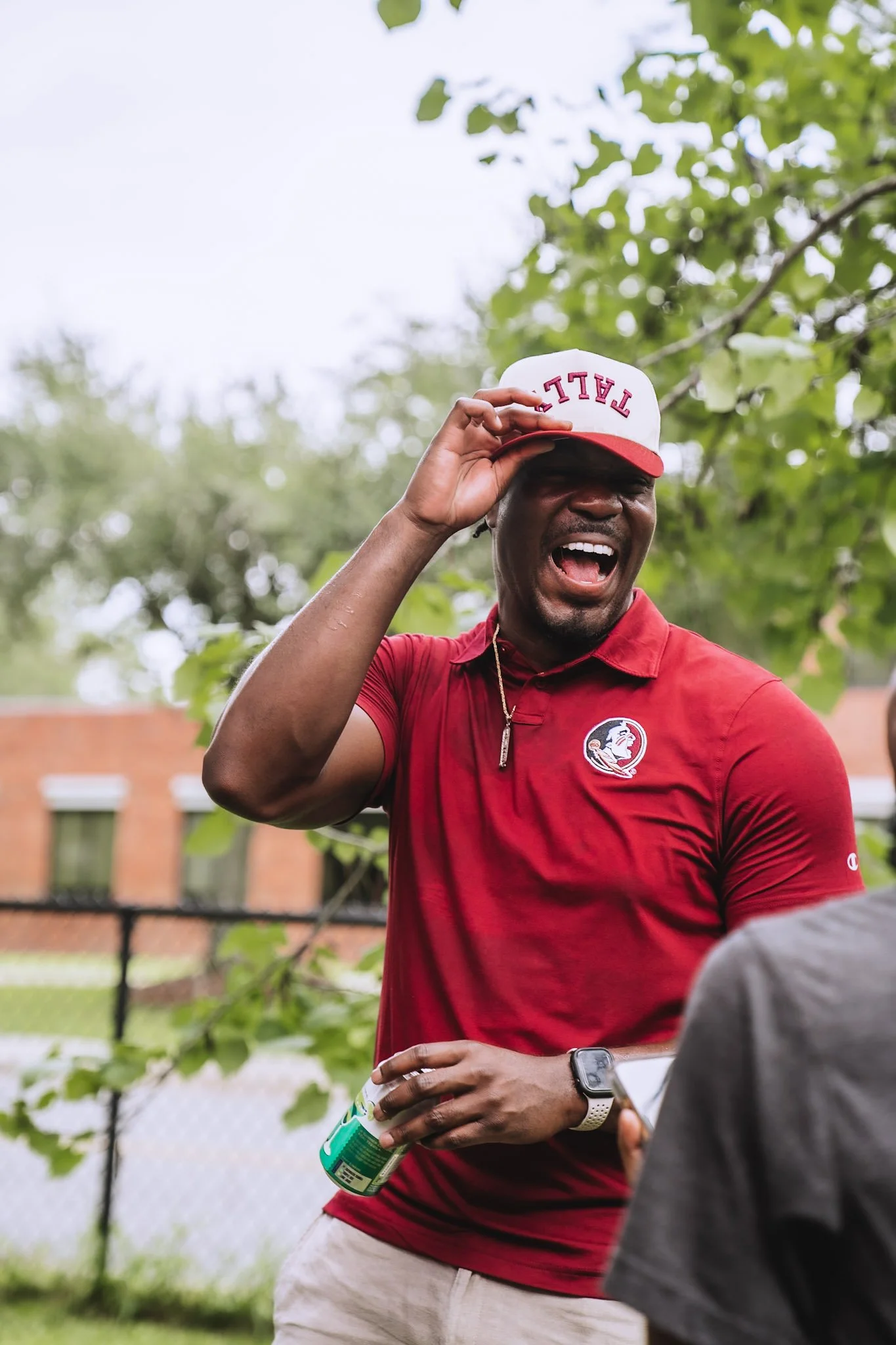 A man wearing a red shirt and a white cap with red lettering laughs and adjusts his cap while holding a green can, standing outdoors near a fenced area with trees in the background.