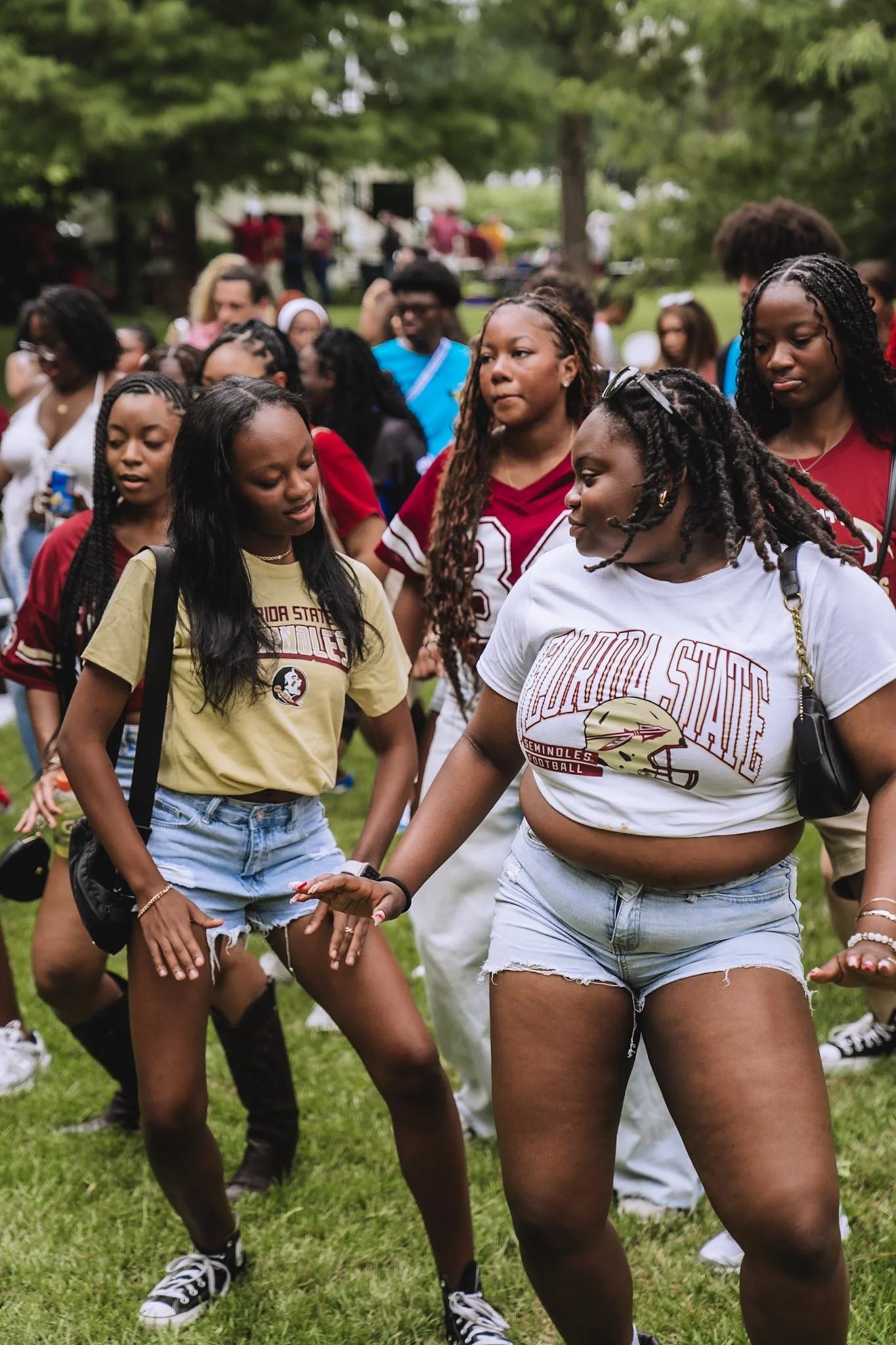 A group of young women and girls gathered outdoors, some dancing or moving to music, with trees and a crowd in the background. Many are wearing Florida State Seminoles football gear.