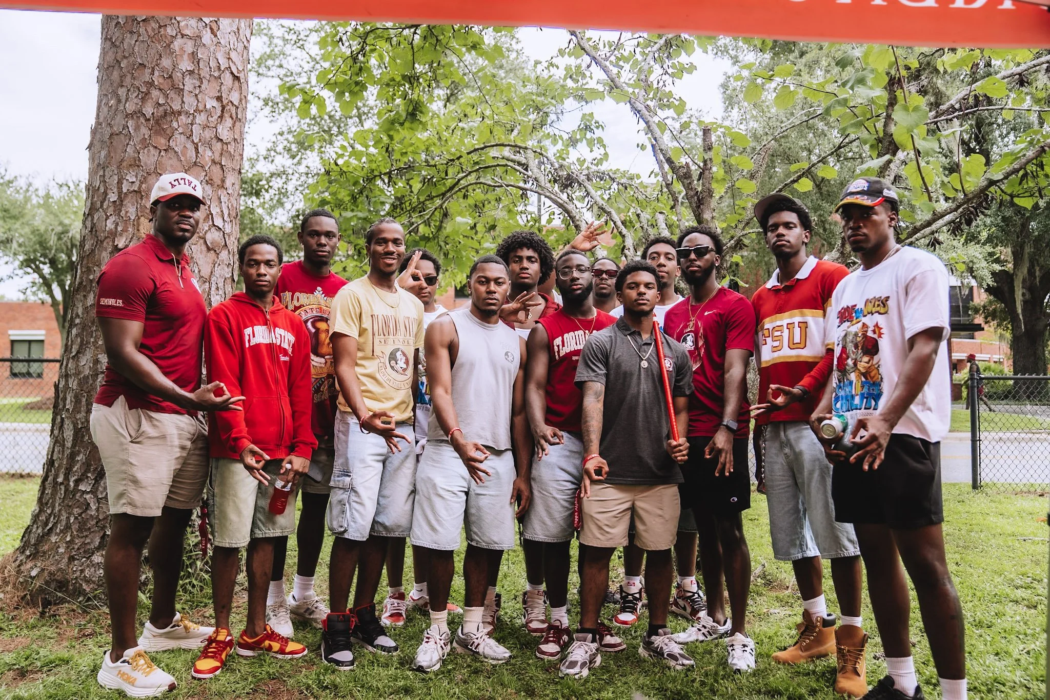 Group of young men posing outdoors in a park, some wearing Florida State University gear, standing near trees and a chain-link fence.