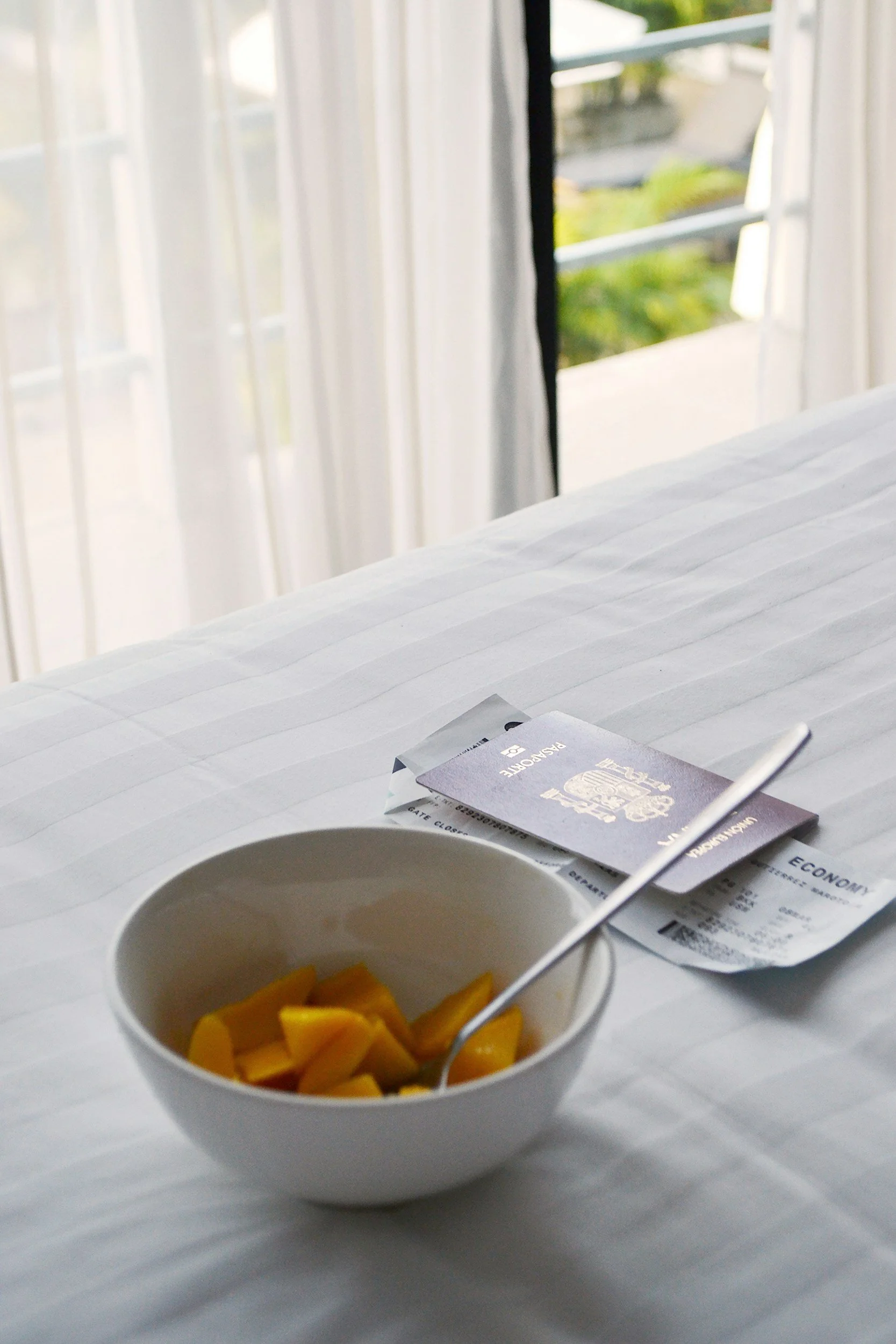 A white bowl filled with chopped mango pieces on a white surface, with a spoon inside. In the background, there are airline tickets and a boarding pass, with a window showing an outdoor view of trees and sky.