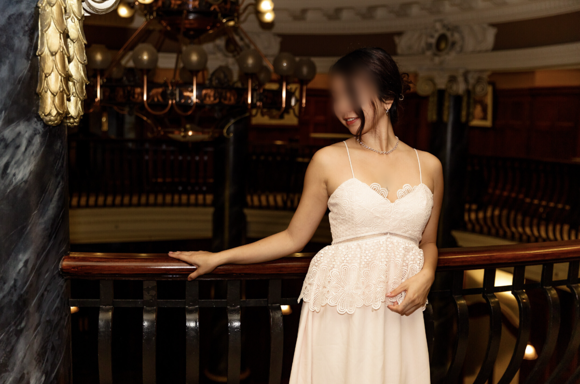 A woman in a white lace dress with spaghetti straps and jewelry, standing by a polished wooden railing in an elegant, dimly lit interior with ornate decor and a chandelier in the background.