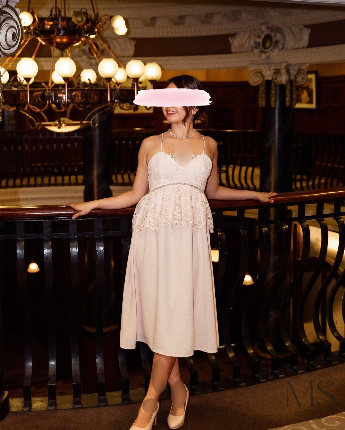 A woman in a cream-colored dress standing with arms resting on a dark wooden railing inside a grand, elegantly decorated building. She is smiling, wearing a necklace and high heels, with a chandelier hanging above in the background.