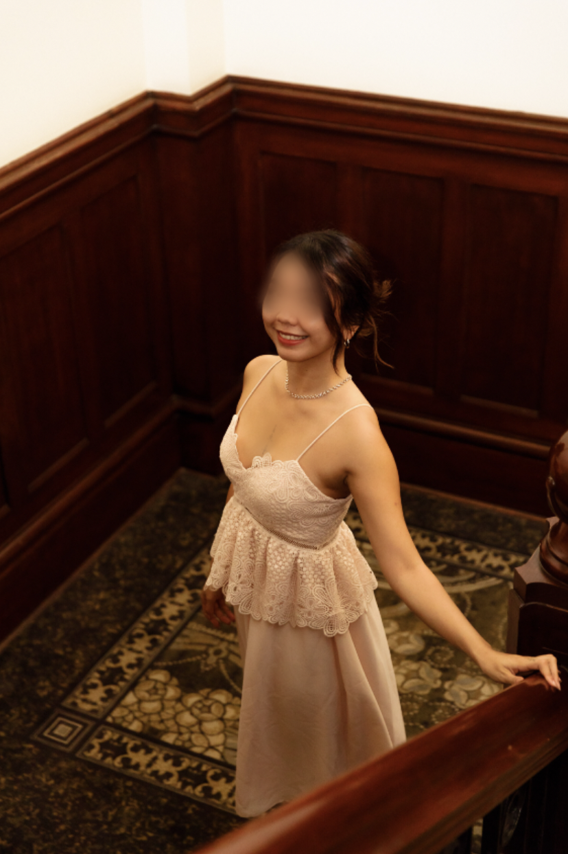 A woman in a pink lace dress standing on a staircase with wood-paneled walls.
