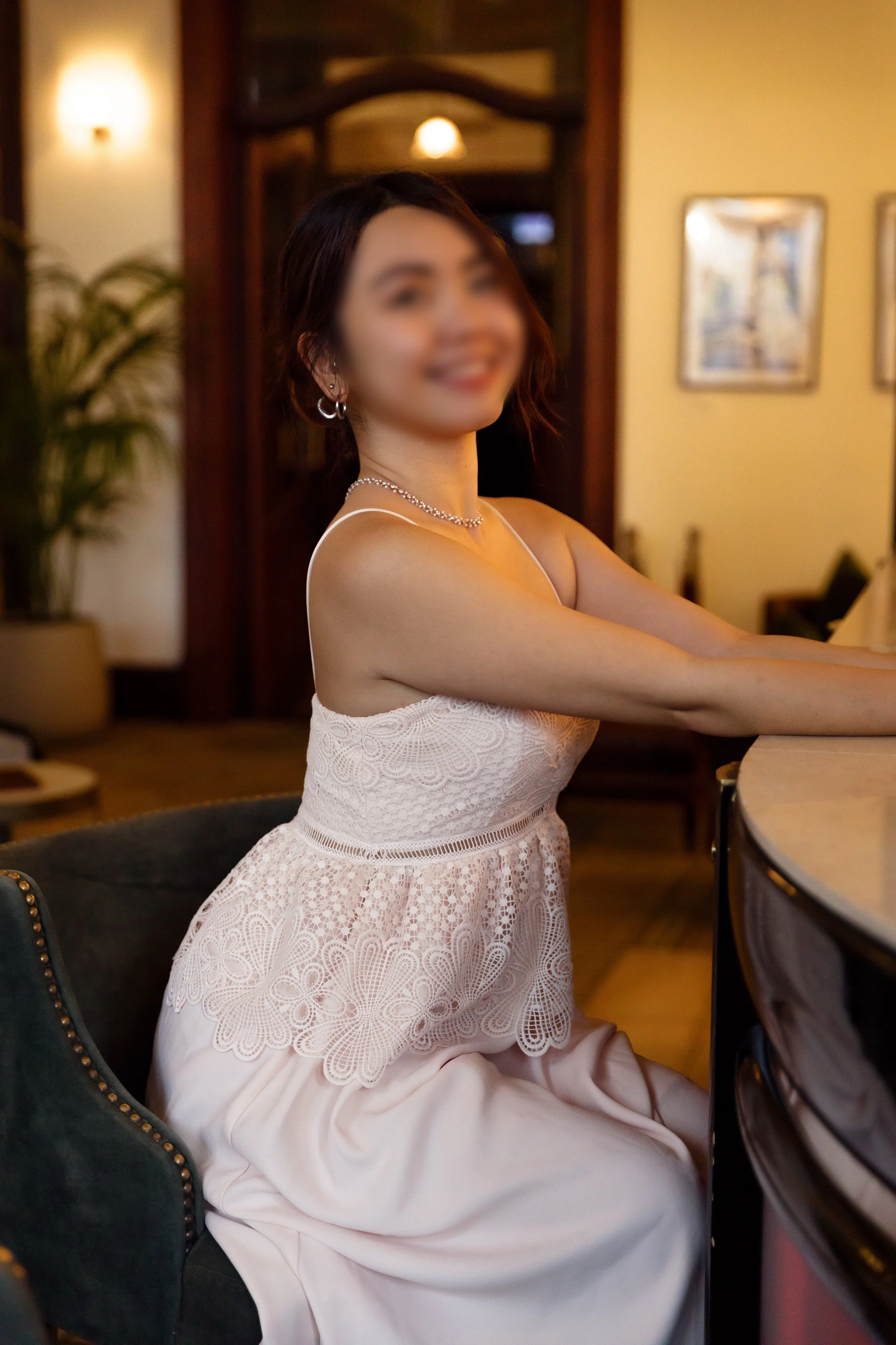 A woman in a white lace dress with pearl jewelry sitting on a green chair at a bar or restaurant, with framed pictures on the wall and warm lighting.