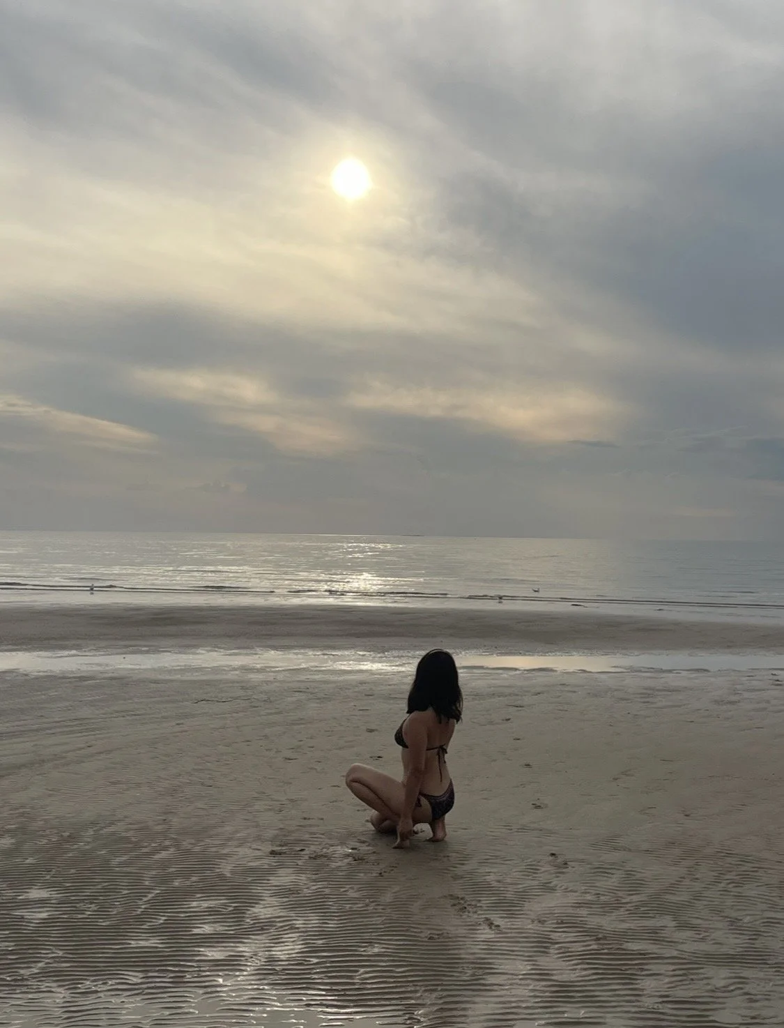 A woman in a swimsuit kneeling on a sandy beach during cloudy weather, with the ocean and a cloudy sky in the background.