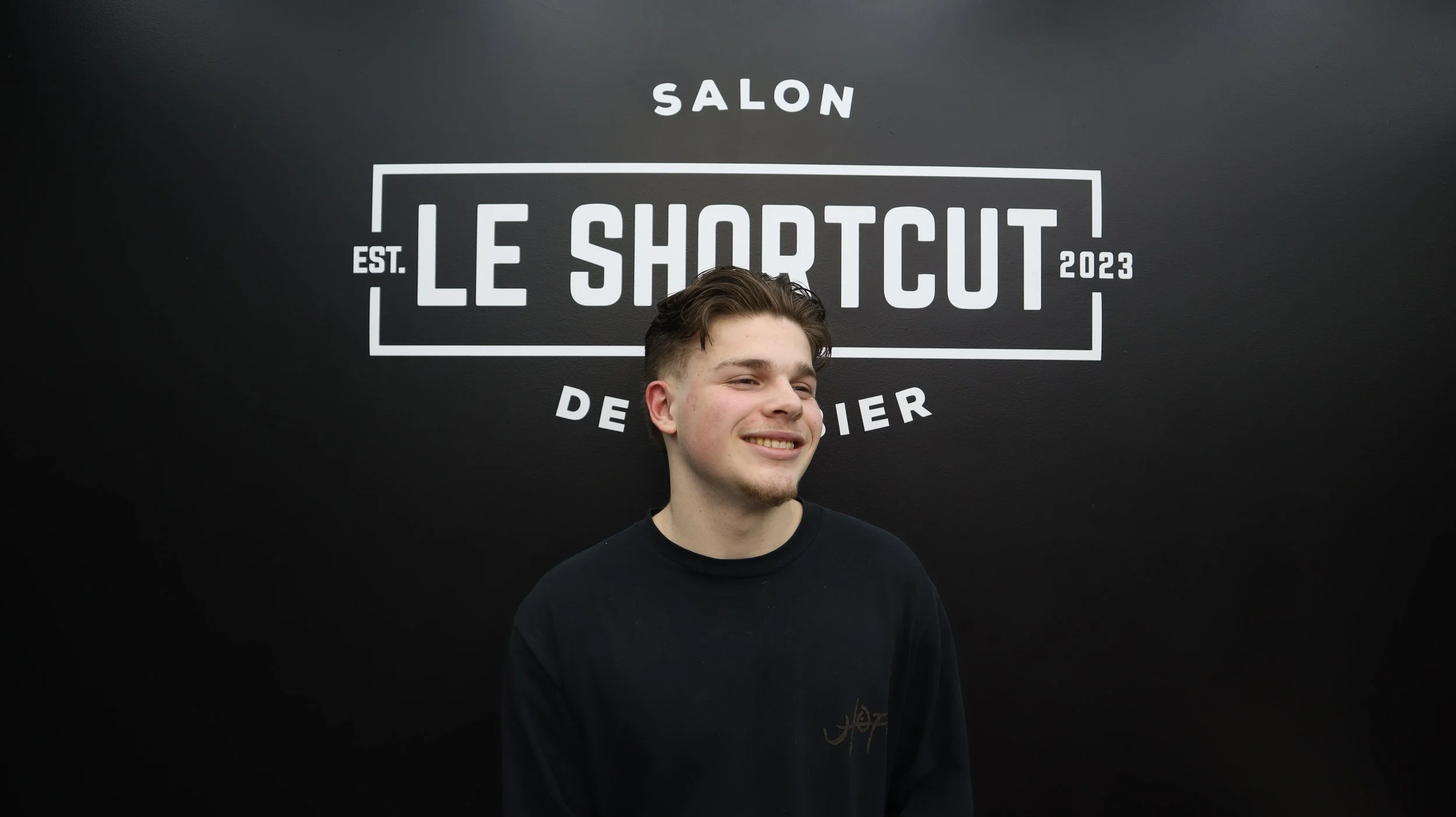 A young man smiling in front of a black wall with the logo of Le Shotcut salon, established in 2023.