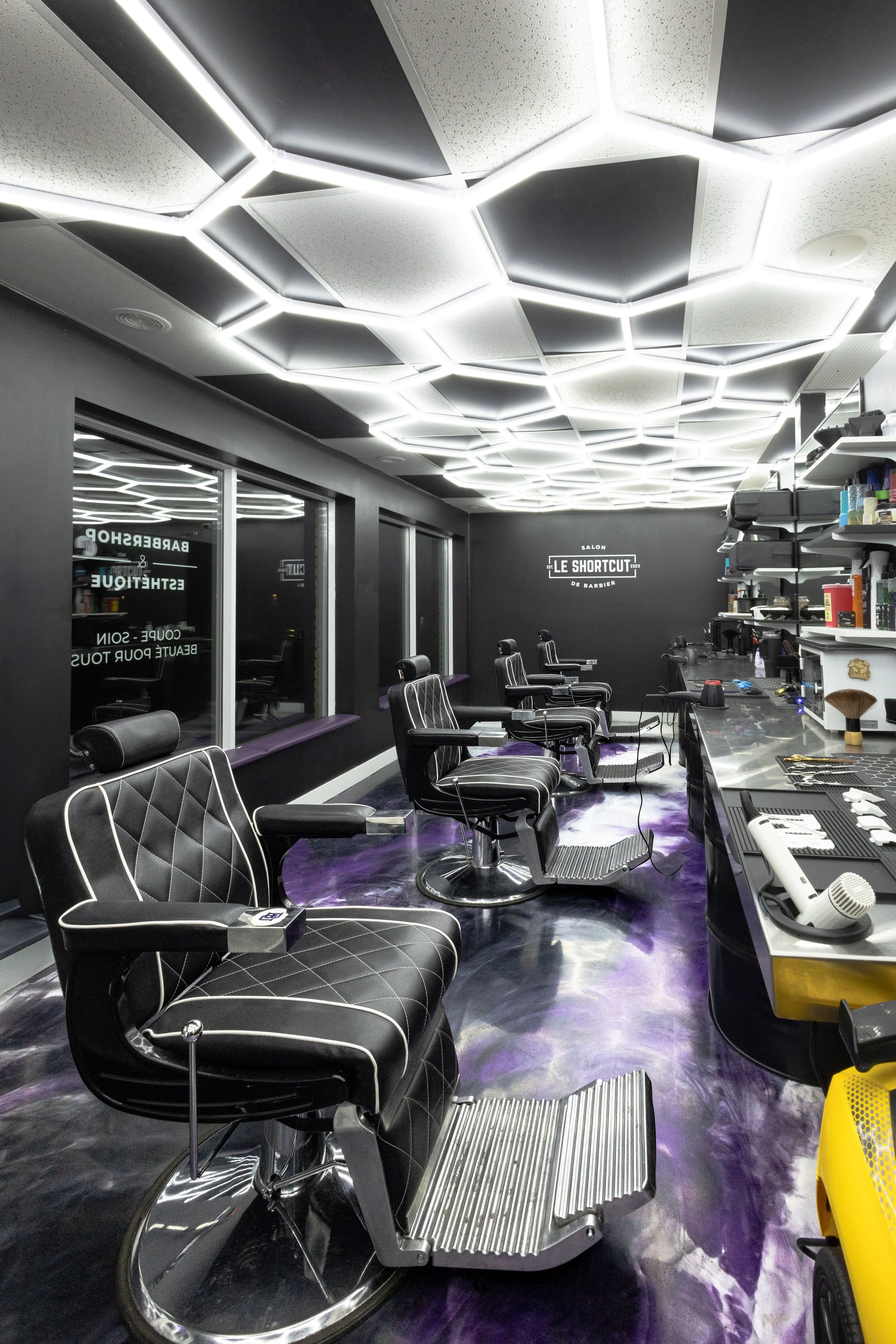 Interior view of a modern barbershop with black and silver barber chairs, a black wall with a sign that reads 'Salon Le Shortcut', and a ceiling with white hexagonal lighting fixtures. There are mirrors, shelves with supplies, and a purple marble-like floor.