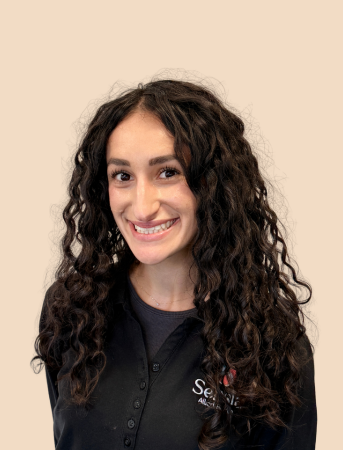 A young woman with curly brown hair smiling at the camera, wearing a black collared shirt with a logo.