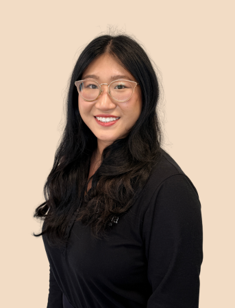 A woman with long dark hair, wearing glasses and a black top, smiling against a beige background.