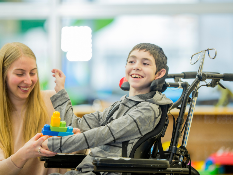 A young boy in a wheelchair smiling and playing with a colorful stacking toy, with a woman beside him in a cheerful environment.