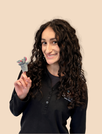 Woman with long curly hair smiling and holding a small gray mouse plush toy in her right hand, against a light beige background.