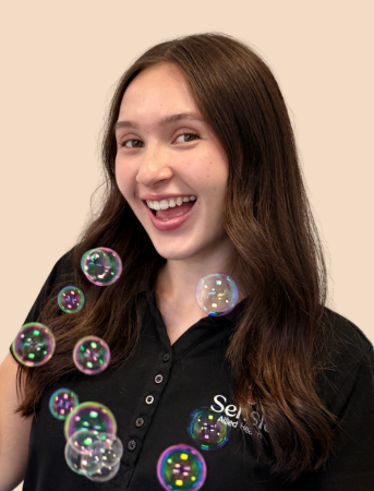 A young woman with long brown hair smiling and looking at a bubble floating in front of her, against a beige background.
