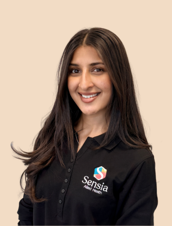 A woman with long dark hair smiling, wearing a black collared shirt with a 'Sensia' logo on it, in front of a beige background.