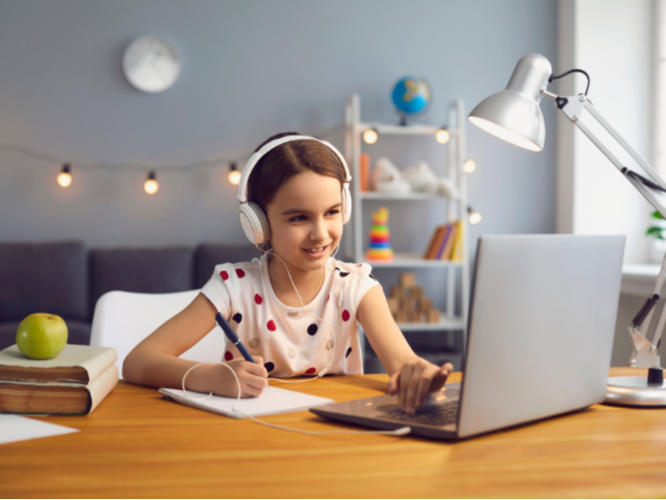 A young girl with headphones smiling while using a laptop at a desk, surrounded by notebooks, an apple, and a desk lamp in a decorated room.