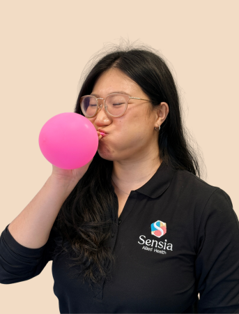 A woman with long dark hair and glasses, wearing a black polo shirt with a Sensia logo, blows up a pink balloon.