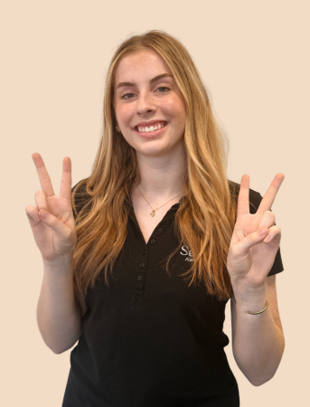 Young woman with long red hair smiling and making peace signs with both hands, wearing a black polo shirt, against a plain beige background.