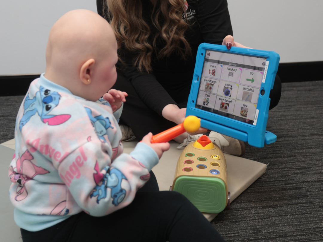 A young child with a bald head and a colorful hoodie playing with a toy hammer while sitting on a mat. An adult, holding a tablet/AAC device with a speech therapy app.
