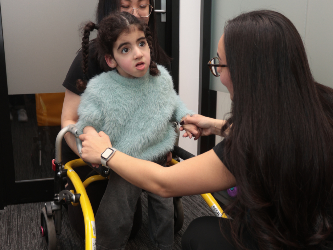 A young girl with curly hair in a blue fuzzy sweater sitting in a yellow wheelchair, talking to a woman with long dark hair and glasses, who is holding her hands.