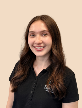 A young woman with long brown hair smiling, wearing a black collared shirt against a beige background.