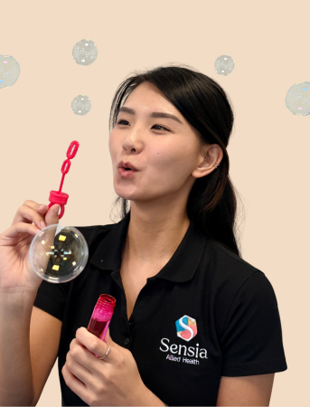 A woman in a black polo shirt blowing bubbles with a bubble wand. She is holding a pink bottle of bubble solution and surrounded by floating bubbles. The shirt has the logo and name 'Sensia Allied Health'.