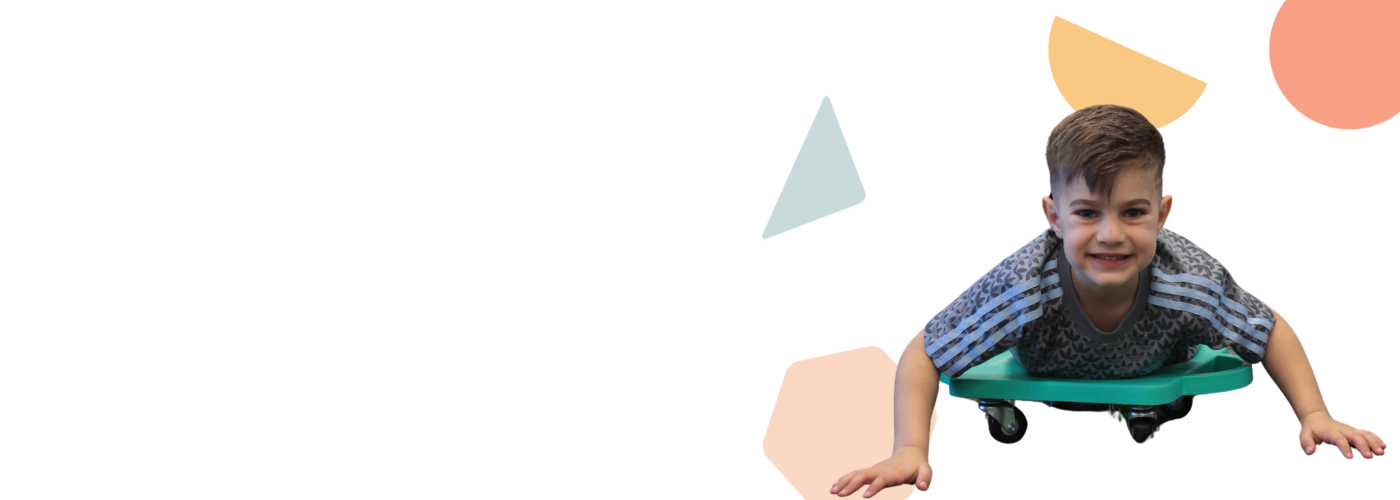 A young boy with short brown hair lying on a green rolling skateboard or tray, smiling at the camera, with a modern, colorful geometric background.