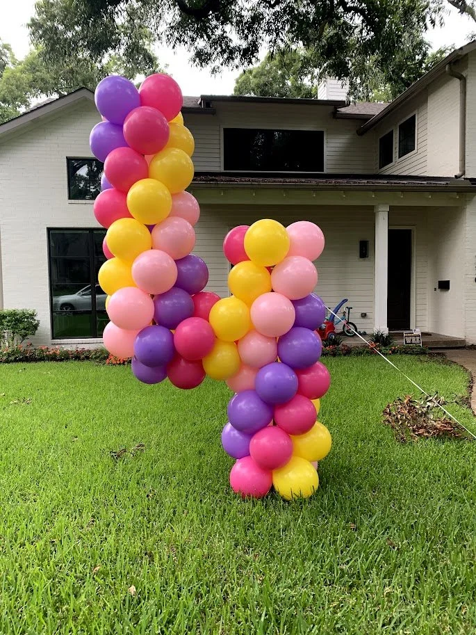 Colorful balloon arrangement in front of a white house with a lawn, consisting of purple, pink, yellow, and red balloons shaped into an abstract form.