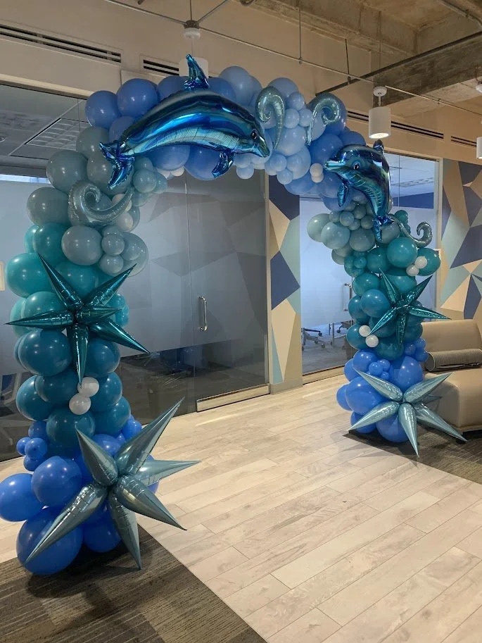 Balloon arch with blue dolphins, star-shaped balloons, and various sizes of blue and white balloons, set up in an indoor space.