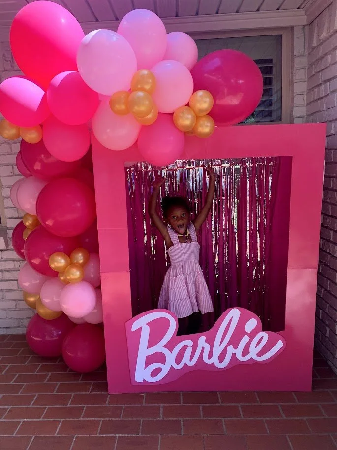 A young girl standing inside a large pink frame with the word 'Barbie' written on it, decorated with pink, red, and gold balloons, against a backdrop of metallic streamers.